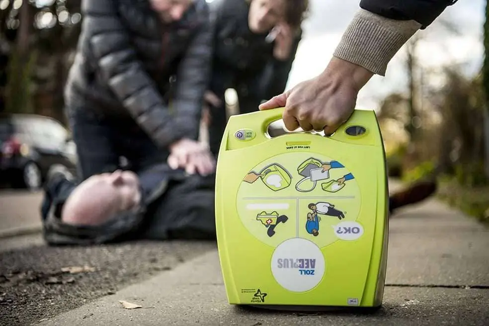 Personer yder hjerte-lunge-redning til en person med hjertestop. En person ankommer på stedet med en hjertestarter.